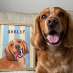 Smiling golden dog named Oakley sitting beside framed custom pet portrait with blue and cream beach stripes background — handmade dog portrait.